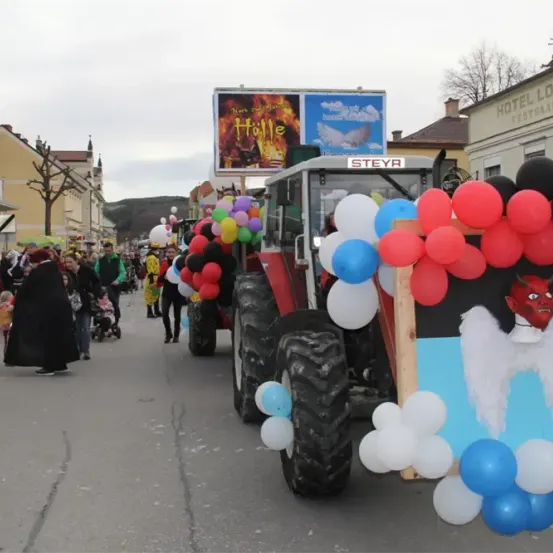 Ein mit Luftballons und einer Teufelsfigur geschmückter Traktor führt einen festlichen Umzug in einer Stadt an, mit Gebäuden und Menschen im Hintergrund.