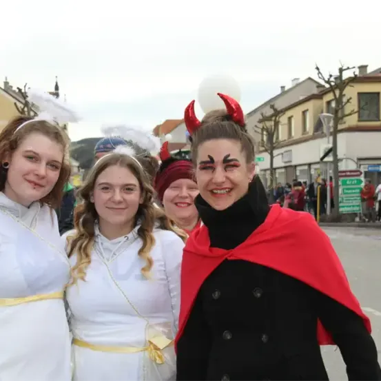 Drei Frauen sind in Kostümen gekleidet und posieren für ein Foto. Zwei tragen weiße Engelkostüme, während die dritte ein rotes Teufelskostüm trägt. Sie stehen in einem Umzug mit verschwommenen Menschen und Gebäuden im Hintergrund.