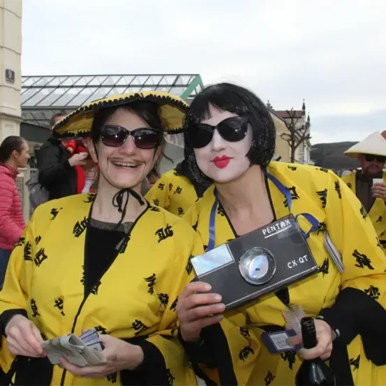 Zwei Frauen in gelben und schwarzen Kostümen posieren für ein Foto mit einer Pentax-Kamera und lächeln in einer Außenumgebung in die Kamera.