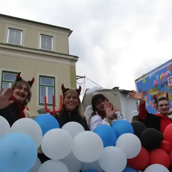 Vier Menschen in Kostümen stehen hinter einem Meer von Luftballons und winken zur Kamera. Dahinter steht ein Gebäude mit Fenstern und einem Banner.