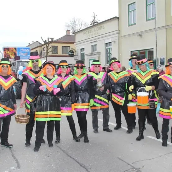 Eine Gruppe von Menschen in bunten Kostümen und mit Eimern steht in einer Parade auf der Straße. Sie tragen Hüte und Brillen. Ein Gebäude mit der Aufschrift Hotel Loibl steht dahinter.
