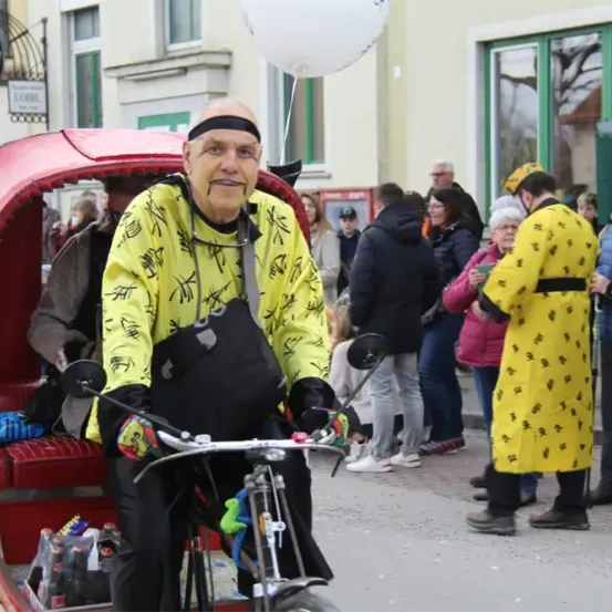 Ein älterer Mann in einem gelben Mantel fährt mit einem roten Wagen ein Fahrrad bei einer öffentlichen Veranstaltung, umgeben von Zuschauern.