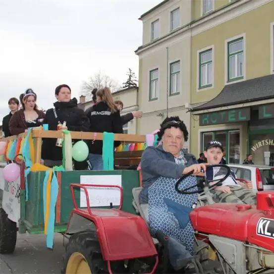 Eine Gruppe von Menschen fährt auf einem dekorierten Traktor durch die Straße. Sie winken und lächeln. Im Hintergrund befindet sich ein Gebäude mit dem Schild Hotel Lois.