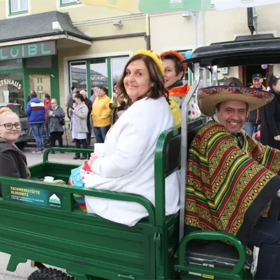 Eine Gruppe von Menschen fährt in einem grünen Wagen bei einer Parade, mit einer Frau in einem weißen Mantel und einem Mann in einem Sombrero. Dahinter stehen Menschen und schauen zu. In der Ferne befindet sich ein Gebäude mit einem Schild, auf dem 'Wirtshaus' steht.
