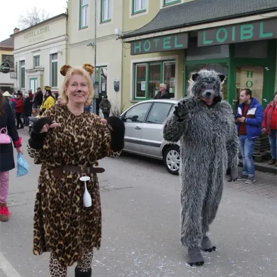 Eine Frau in einem Leoparden-Kostüm und ein Mann in einem Bären-Kostüm gehen auf einer Straße, hinter ihnen steht eine Menge. Vor ihnen steht ein silbernes Auto, das vor einem Gebäude mit dem Wort Hotel geparkt ist.