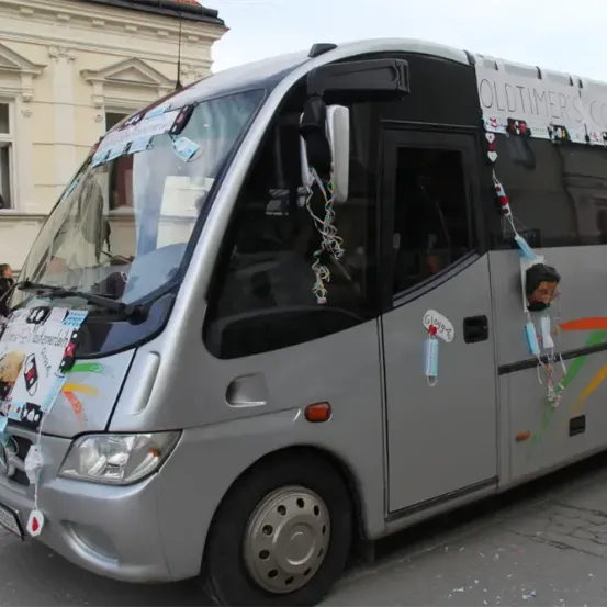 Ein silberner Bus mit Dekorationen und einem Schild, auf dem Oldtimer's Cordon steht. Die Dekorationen bestehen aus bunten Bändern, Aufklebern und einer Comicfigur. Er ist vor einem Gebäude mit Fenster geparkt.