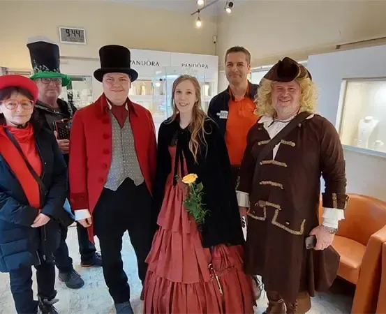 Sechs Personen stehen in einem Raum, gekleidet in Kostüme. Eine Frau in einem roten Kleid hält eine Blume. Hinter ihnen hält ein Mann ein Gerät. Im Hintergrund sind Displays und Mannequins zu sehen.