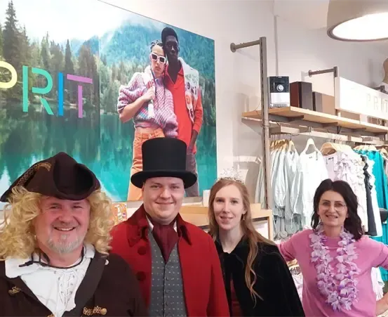 Vier Menschen in Kostümen stehen vor einem Bekleidungsgeschäft. Ein großes Schild mit dem Wort Spirit ist hinter ihnen.