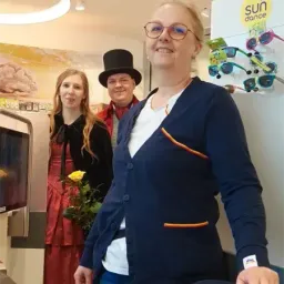 Drei Personen stehen in einem Raum. Eine Frau in der Mitte hält eine Blume, während ein Mann links einen roten Mantel und Hut trägt. Hinter ihnen sind eine Mikrowelle und ein Schaukasten mit dem Text 'sun dance' zu sehen.