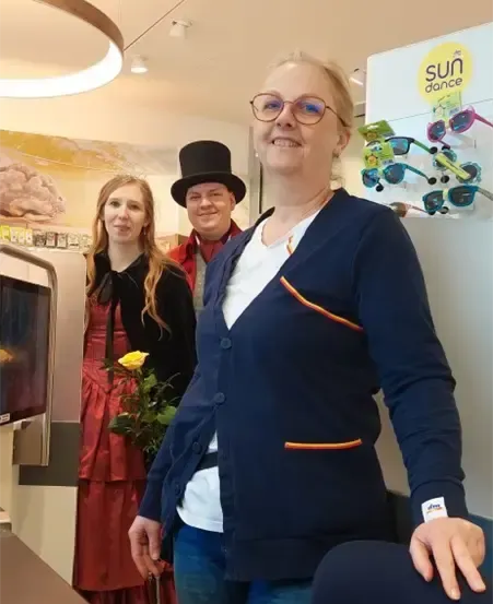 Drei Personen stehen in einem Raum. Eine Frau in der Mitte hält eine Blume, während ein Mann links einen roten Mantel und Hut trägt. Hinter ihnen sind eine Mikrowelle und ein Schaukasten mit dem Text 'sun dance' zu sehen.