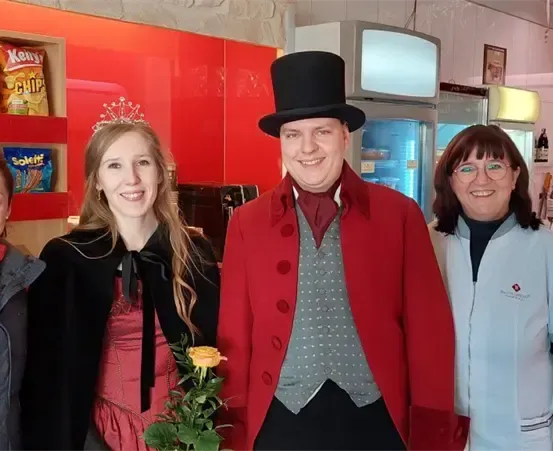Vier Personen stehen in einem weihnachtlich dekorierten Laden. Ein Mann in einem roten Mantel und einem Zylinder hält eine gelbe Rose. Die Frau neben ihm trägt ein rotes Kleid. Hinter ihnen befindet sich ein roter Regal mit Snacks und einem Kühlschrank.