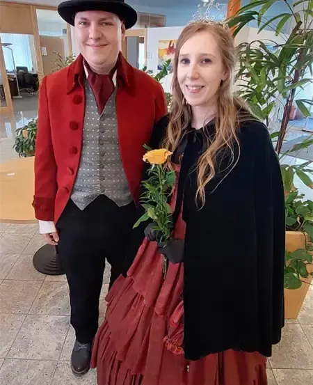 Ein Mann und eine Frau in historischer Kleidung, lächelnd und posierend für ein Foto. Der Mann trägt einen roten Mantel und einen hohen Hut, während die Frau ein rotes Kleid und einen schwarzen Umhang trägt.