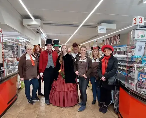 Eine Gruppe von Menschen in verschiedenen Kostümen, darunter ein viktorianisches Kleid, einen hohen Hut und eine rote Haube, posiert für ein Foto in einem Geschäft.