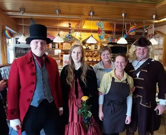 Eine Gruppe von Menschen in Kostümen posiert für ein Foto in einer Bäckerei mit Dekorationen, die von der Decke hängen.