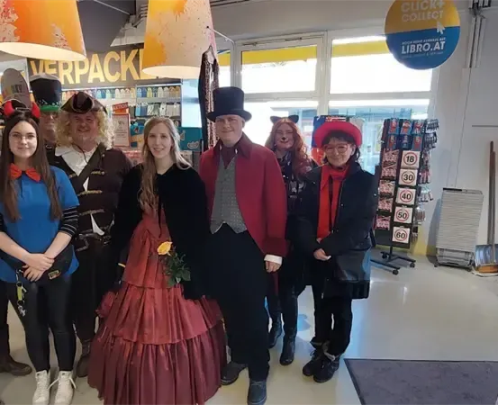 Eine Gruppe von Menschen in verschiedenen Kostümen steht in einem Geschäft. Eine Frau hält eine Blume, und andere tragen Hüte und Brillen. Hinter ihnen sind Regale mit Produkten und ein gelbes Schild mit dem Wort 'Verpack' zu sehen.