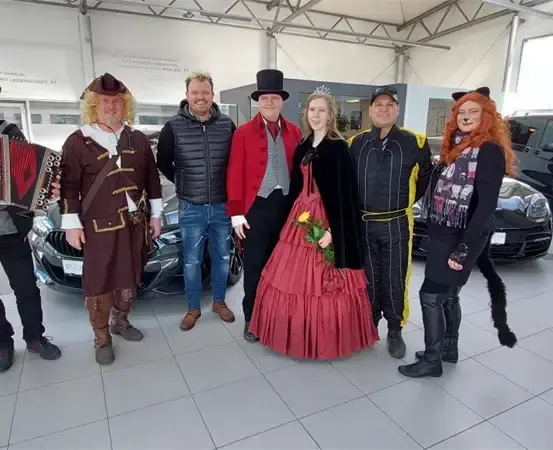 Eine Gruppe von Menschen in Kostümen steht in einem Autohaus, mit Autos und einer weißen Wand im Hintergrund.