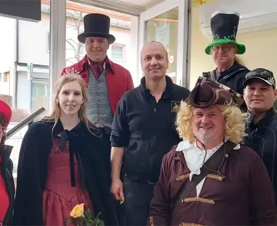 Eine Gruppe von Menschen in Kostümen, einschließlich Hüten und Kleidern, posiert für ein Foto. Dahinter befindet sich ein Gebäude mit Fenstern und einer Treppe.