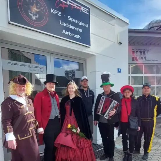 Eine Gruppe von Menschen in historischen Kostümen steht vor einem Gebäude mit dem Schild 'KFZ - Lackiererei Airbrush'. Die Frau in der Mitte hält einen Blumenstrauß.