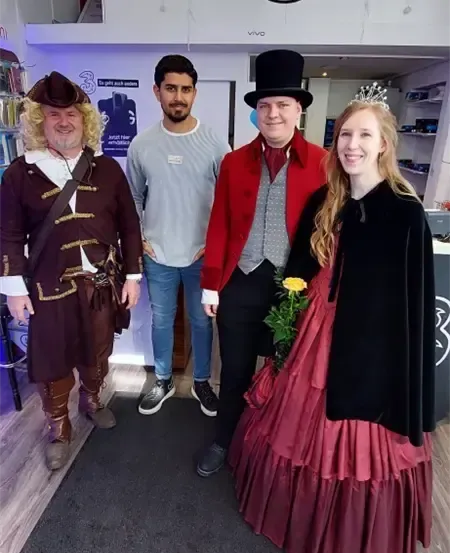 Vier Personen in Kostümen posieren für ein Foto in einem Raum. Ein Mann in Piratenkostüm steht links, während eine Frau in viktorianischem Kleid rechts steht. Dazwischen stehen zwei Männer in formeller Kleidung.