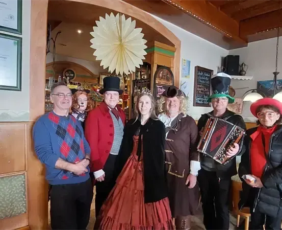 Eine Gruppe von Menschen in Kostümen, darunter eine Frau mit einem Akkordeon, posiert für ein Foto in einem dekorierten Raum mit Lampen und gerahmten Bildern an der Wand.