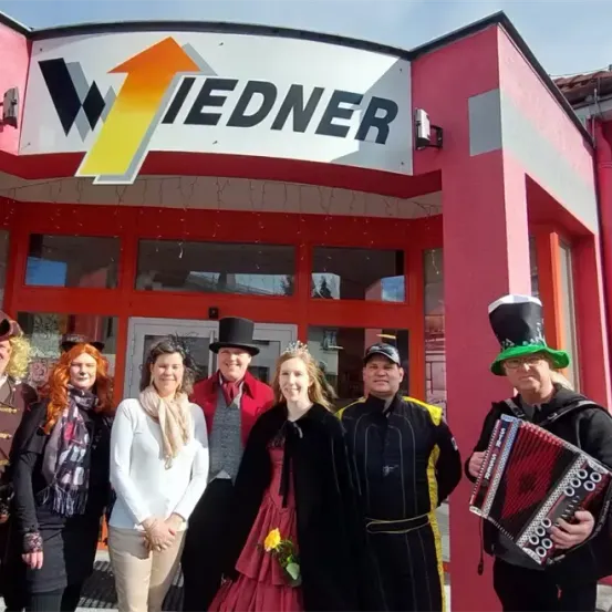 Eine Gruppe von Menschen in Kostümen steht vor einem Gebäude mit einem roten Schild, auf dem Wiedner steht. Die Kostüme variieren, darunter ein Pirat, eine Prinzessin und ein Mann mit einem Akkordeon.