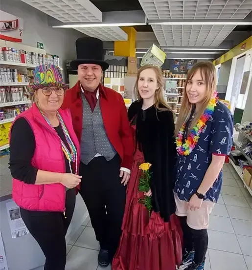 Vier Menschen stehen in einem Laden, zwei Frauen und zwei Männer. Sie sind in Kostümen gekleidet.