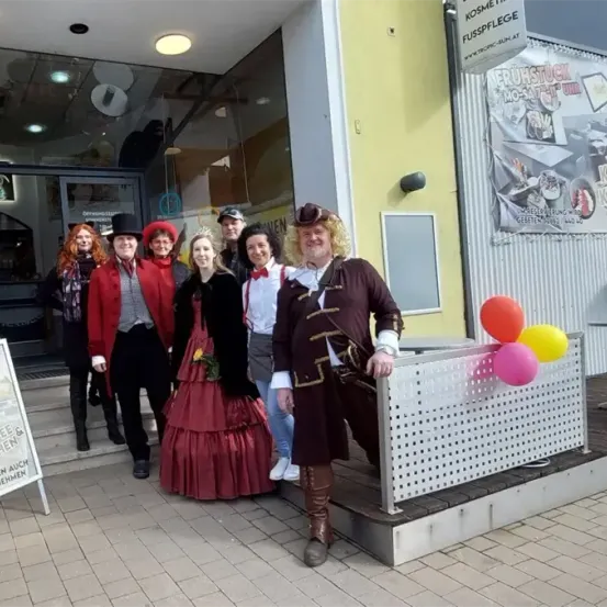 Eine Gruppe von Menschen in Kostümen steht vor einem Geschäft. Sie lächeln und scheinen für ein Foto zu posieren.