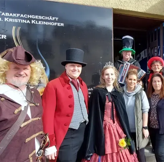 Eine Gruppe von Menschen in Kostümen steht vor einem Ladenschild mit der Aufschrift 'Tabakfachgeschaft Inh. Kristina Kleinhofer'. Sie scheinen für ein Foto zu posieren.