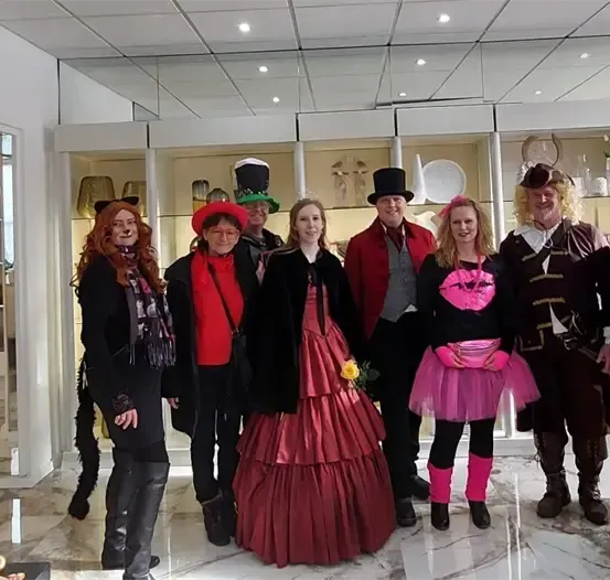 Eine Gruppe von Menschen in Kostümen, darunter ein rotes Kleid und Hüte, posiert für ein Foto in einem Raum mit Regalen und weißen Wänden.