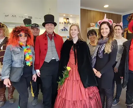 Eine Gruppe von Menschen in Kostümen posiert für ein Foto, einige tragen Hüte und Brillen. Eine Frau in einem rosa Kleid steht in der Mitte.