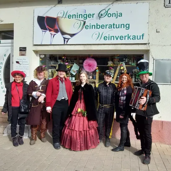 Sieben Menschen in Kostümen stehen vor einem Weinladen. Das Schild des Ladens lautet 'Weninger Sonja Weinberatung Weinverkauf'. Sie lächeln für die Kamera.