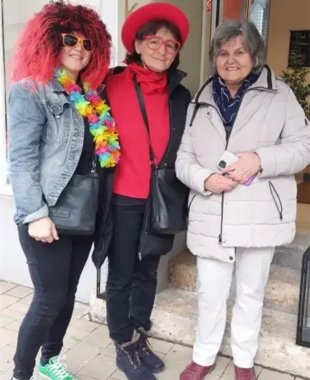 Drei Frauen stehen lächelnd vor dem Eingang eines Ladens. Die Frau links trägt eine rote Perücke und eine bunte Girlande. Die Frau in der Mitte trägt einen roten Hut und eine Brille. Die Frau rechts hält eine Kamera.