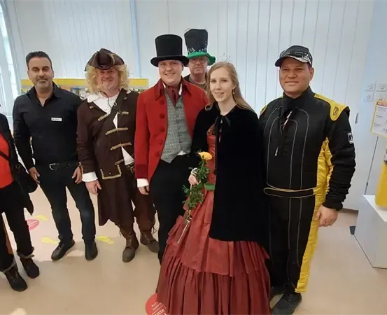 Eine Gruppe von Menschen in verschiedenen Kostümen steht zusammen, lächelt und posiert für ein Foto. Ein Mann trägt einen roten Mantel, ein anderer einen schwarzen Rennanzug, und eine Frau hält eine Blume.