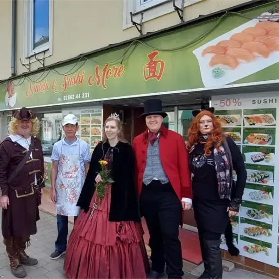 Eine Gruppe von Menschen in Vintage-Kostümen steht vor einem Restaurant. Die Speisekarte bietet Sushi mit 50% Rabatt.