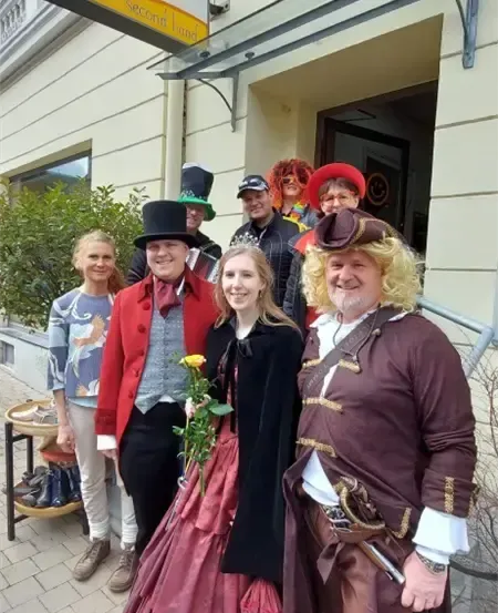 Eine Gruppe von Menschen in verschiedenen Kostümen, einschließlich Hüten und Stiefeln, steht vor einem Geschäft mit einem Schild, das 'Der Trend Second Hand' lautet. Sie lächeln und posieren für ein Foto.