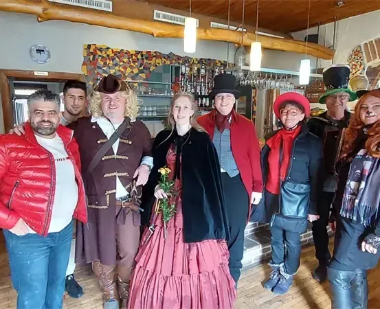 Eine Gruppe von Menschen in verschiedenen Kostümen, einschließlich Hüten und Handschuhen, steht in einem Raum mit Holzböden und Regalen voller Flaschen.