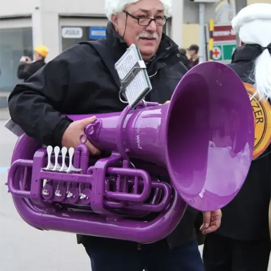 Ein Mann mit weißem Hut und Brille hält eine lila Tuba in einer Außenumgebung, möglicherweise bei einer Parade, mit anderen Personen im Hintergrund.