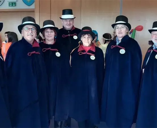 Eine Gruppe von Erwachsenen in passenden blauen Umhängen und Hüten, mit einer Frau vorne in einem roten Umhang, lächelt für ein Foto. Dahinter ist eine Tür mit einem Schild und einem Zeichen zu sehen.