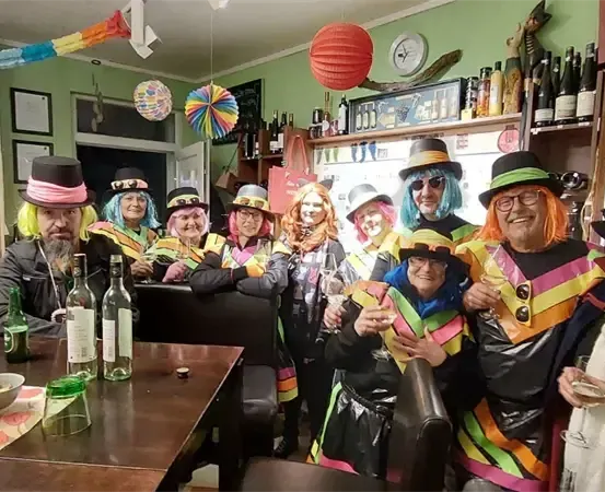 Eine Gruppe von Menschen in bunten Kostümen, einige mit Gläsern, versammelt sich in einem Raum mit dekorierter Wand und einem Regal mit Flaschen.