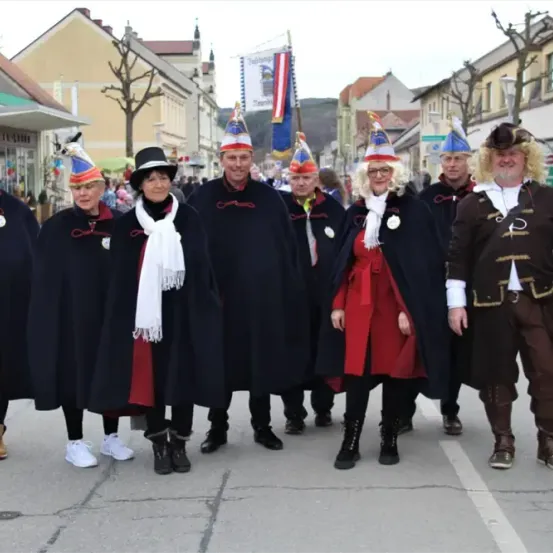 Eine Gruppe von Menschen in Kostümen steht auf einer Straße. Einige tragen Hüte. Die Frau in Rot hat eine Brille. Sie posieren für ein Foto. Dahinter stehen Gebäude und Bäume.