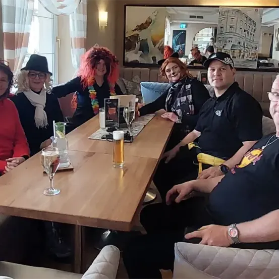 Eine Gruppe von sechs Personen sitzt um einen Holztisch mit Getränken und einem Menü und lächelt für ein Foto. Der Hintergrund zeigt einen Spiegel, der Menschen und ein Wandbild reflektiert.