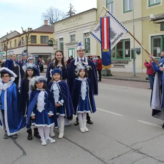 Eine Gruppe von Menschen in blauen Kostümen, möglicherweise für eine Parade, geht auf der Straße. Sie halten eine Flagge. Am Straßenrand stehen Gebäude und Bäume.