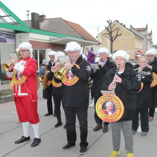 Eine Gruppe von Musikern in Kostümen spielt Instrumente bei einem Umzug. Dazu gehören Trompeten, Posaunen und eine Klarinette. Sie stehen vor einem Gebäude mit einem Schild, auf dem Blue steht.