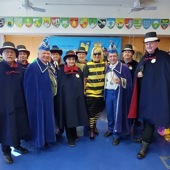 Eine Gruppe von Erwachsenen in Kostümen posiert für ein Foto in einem Raum mit blauem Boden und einem Banner im Hintergrund.