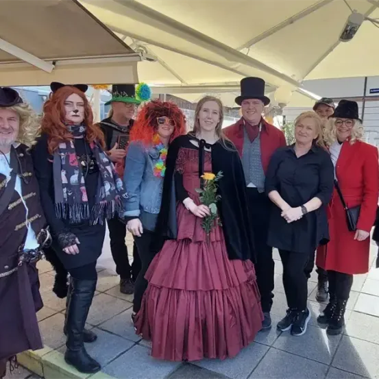 Eine Gruppe von Menschen in Kostümen und Hüten steht unter einem Schirm auf einem Bürgersteig. Eine Frau in einem roten Kleid hält eine Blume.