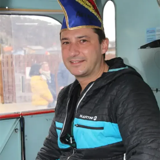 Ein Mann in einer schwarzen und blauen Jacke mit einem blauen Hut sitzt auf einem Fahrzeug und lächelt in die Kamera. Im Hintergrund ist eine verschwommene Ansicht einer weiteren Person und eines Zauns zu sehen.