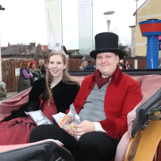 Ein Mann und eine Frau in Vintage-Kleidung sitzen in einer Kutsche und lächeln. Sie halten Süßigkeiten und sitzen gegen einen rosa Sitz. Dahinter ein Gebäude mit einem Schild Nummer 11 und Zuschauer.