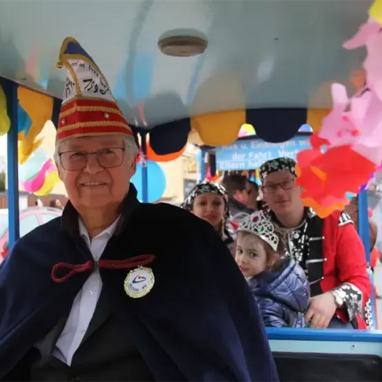 Ein älterer Mann mit einem traditionellen Hut fährt in einer geschmückten Kutsche mit Kindern und Erwachsenen. Die Kutsche hat bunte Dekorationen und Luftballons. Im Hintergrund sind ein rotes Auto und ein Gebäude zu sehen.