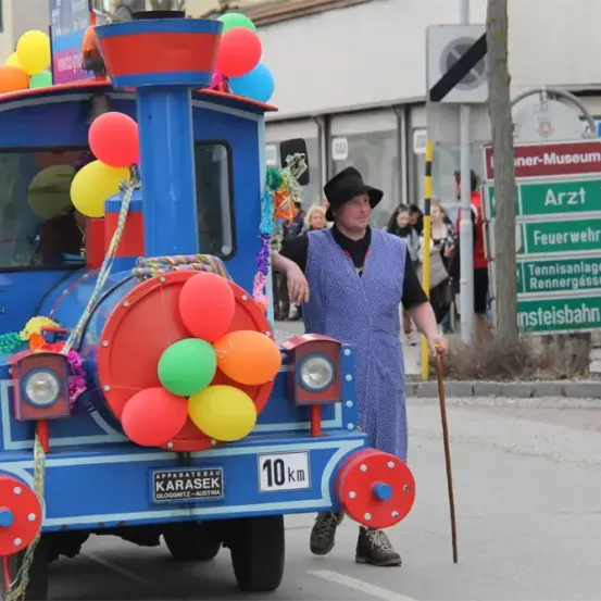Ein Mann in einem blauen Kleid geht neben einem dekorierten blauen Zug auf der Straße. Straßenschilder und Ballons sind vorhanden.