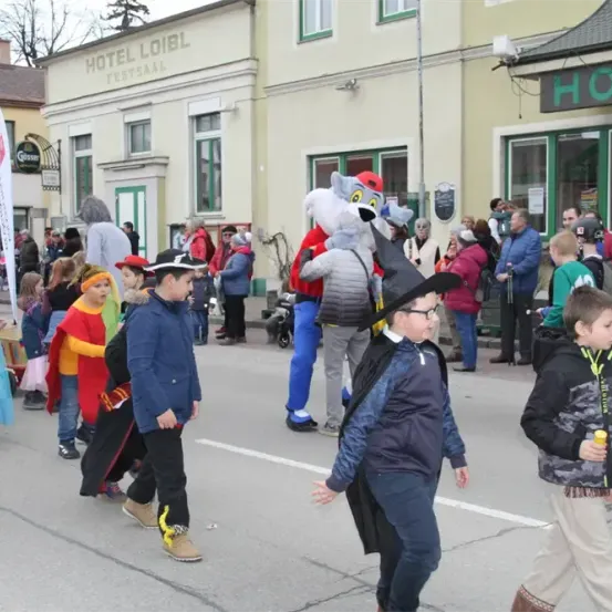 Ein Umzug mit Menschen in verschiedenen Kostümen, darunter eine Person in einem Hundekostüm, die mitten auf der Straße geht. Auf beiden Seiten der Straße befinden sich Gebäude.
